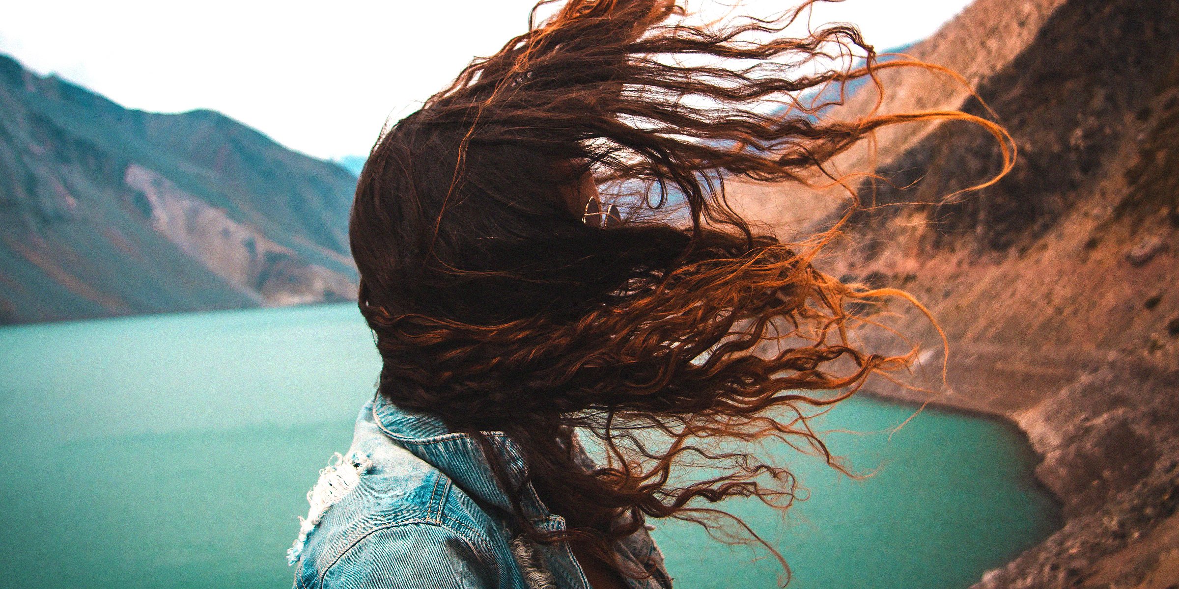 Unsplash | A woman with her red hair blowing in the wind as the stands in front of scenery filled with mountains and a river