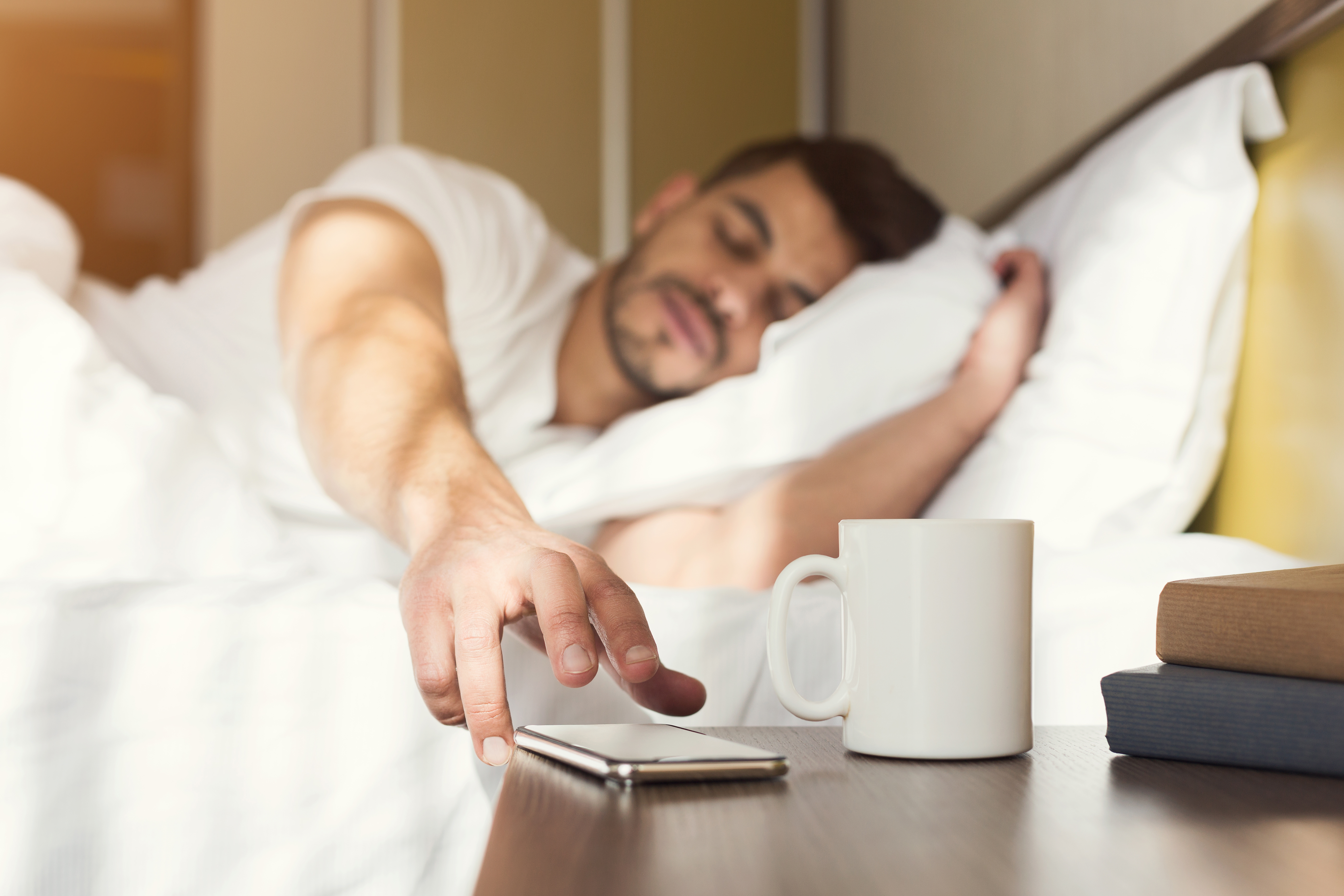 Chico somnoliento que se despierta temprano tras oír la señal del despertador en el smartphone. | Fuente: Shutterstock
