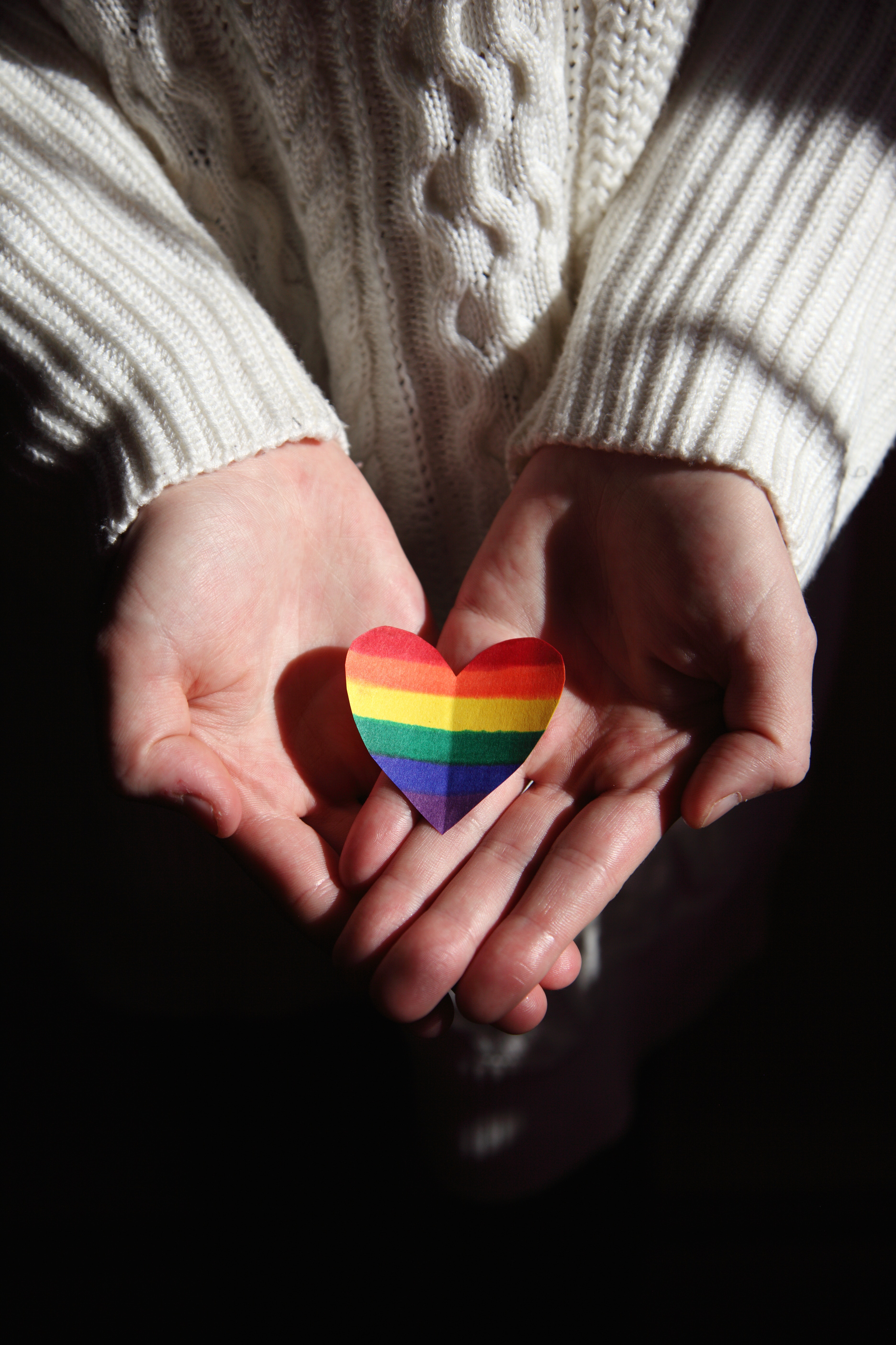 An individual cupping a cut out heart with the Pride colors on it. │ Source: Unsplash