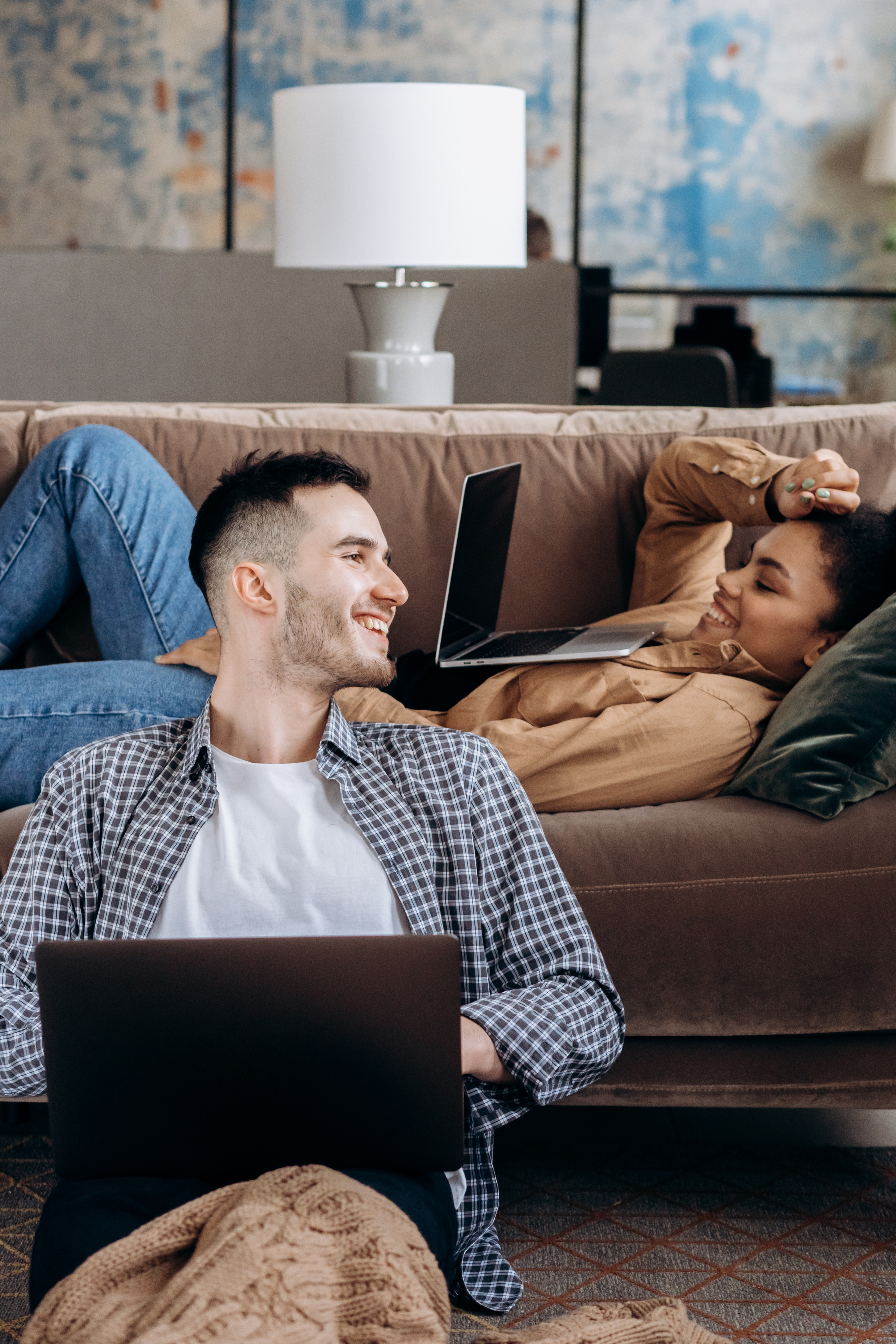 Man In Black And White Checkered Dress Shirt Sitting on Floor With A Woman Lying On A Couch Both Using Laptops. | Source: Pexels