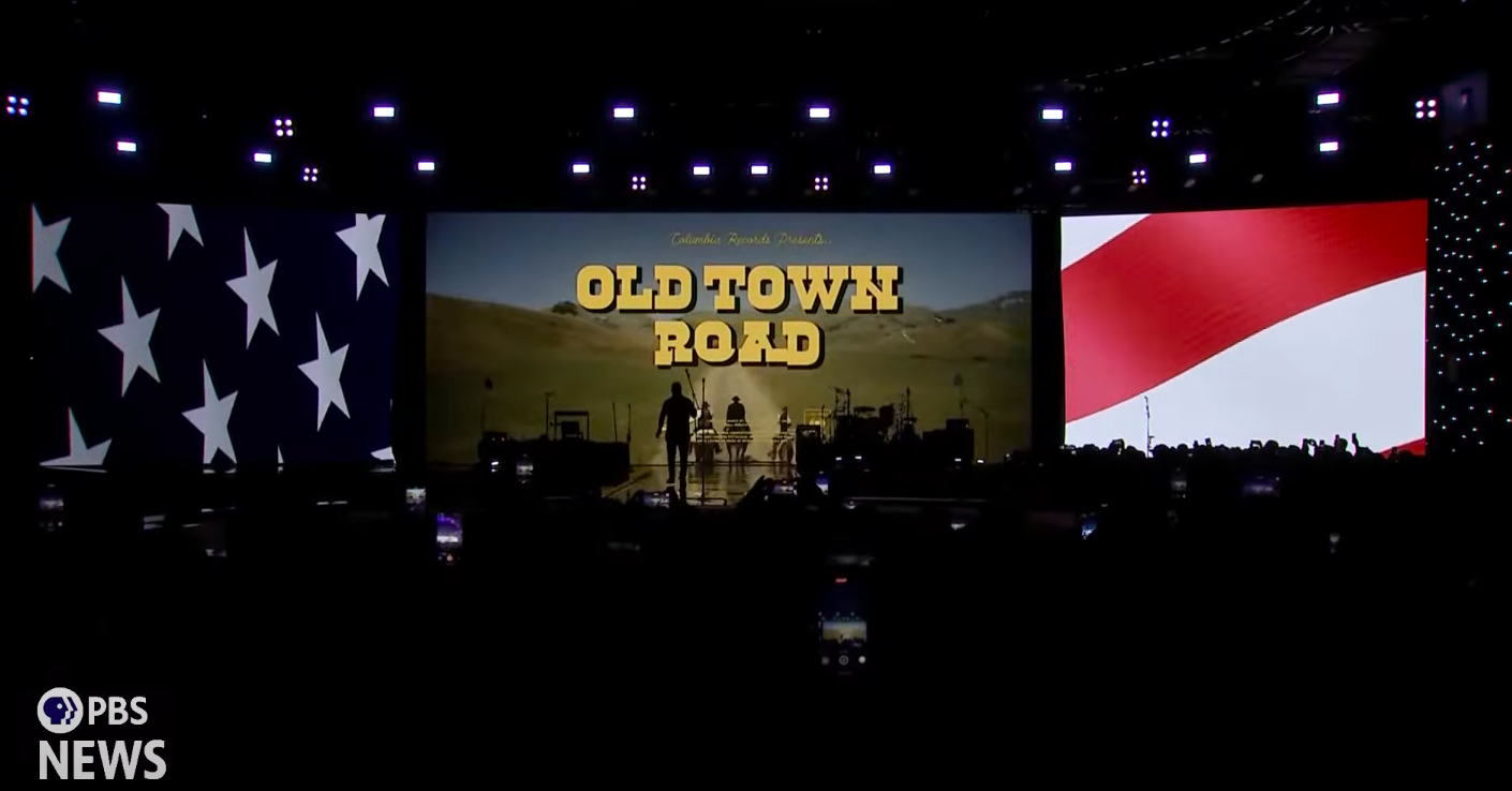 The "Old Town Road" music video playing on a projector screen on stage right before Billy Ray Cyrus appears for his performance during the Liberty Ball, posted on January 21, 2025. | Source: YouTube: PBS NewsHour